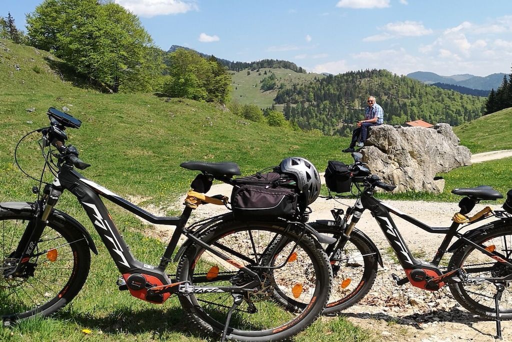 Two bicycles with attached gear bags and helmets, parked on a grassy path with a hilly landscape in the background.
