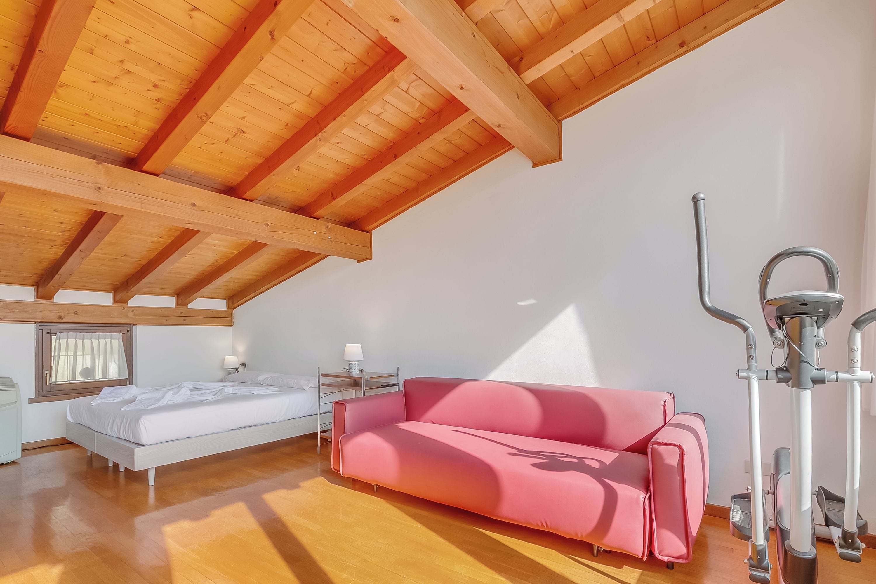 A room with a wooden ceiling, a bed, a pink sofa, and exercise equipment.