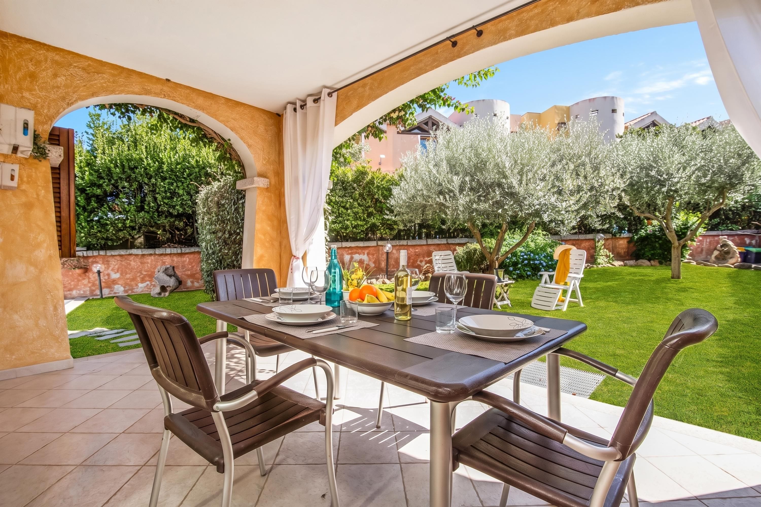 An outdoor patio with a table set for dining, surrounded by chairs, and a view of a well-maintained garden and buildings in the background.