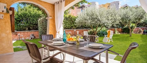 An outdoor patio with a table set for dining, surrounded by chairs, and a view of a well-maintained garden and buildings in the background.