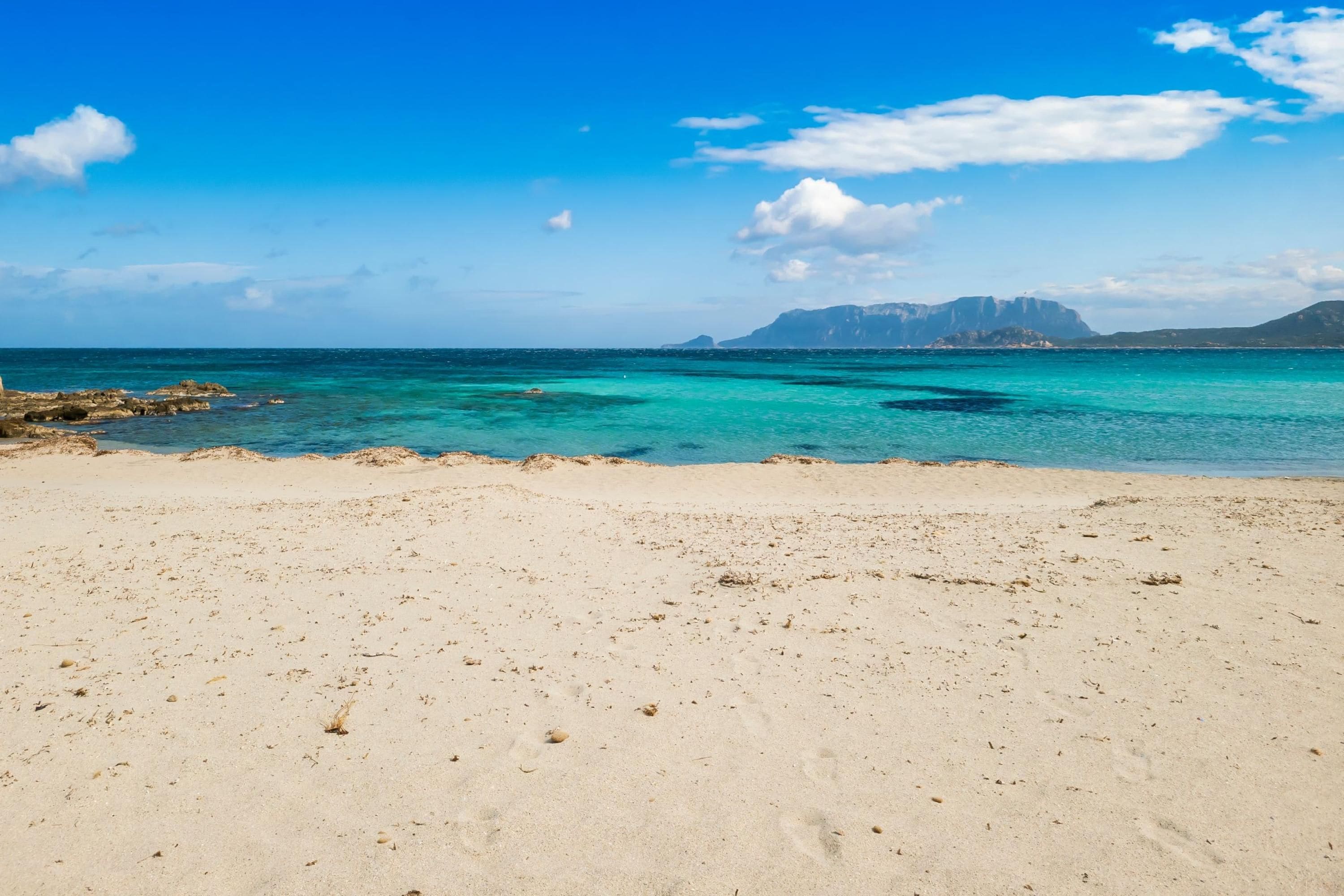 A beach with clear turquoise water and distant mountains under a blue sky.