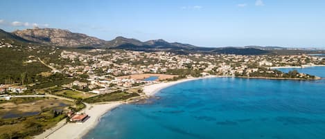 An aerial view of a coastal town with a beach, clear blue waters, and mountainous background.
