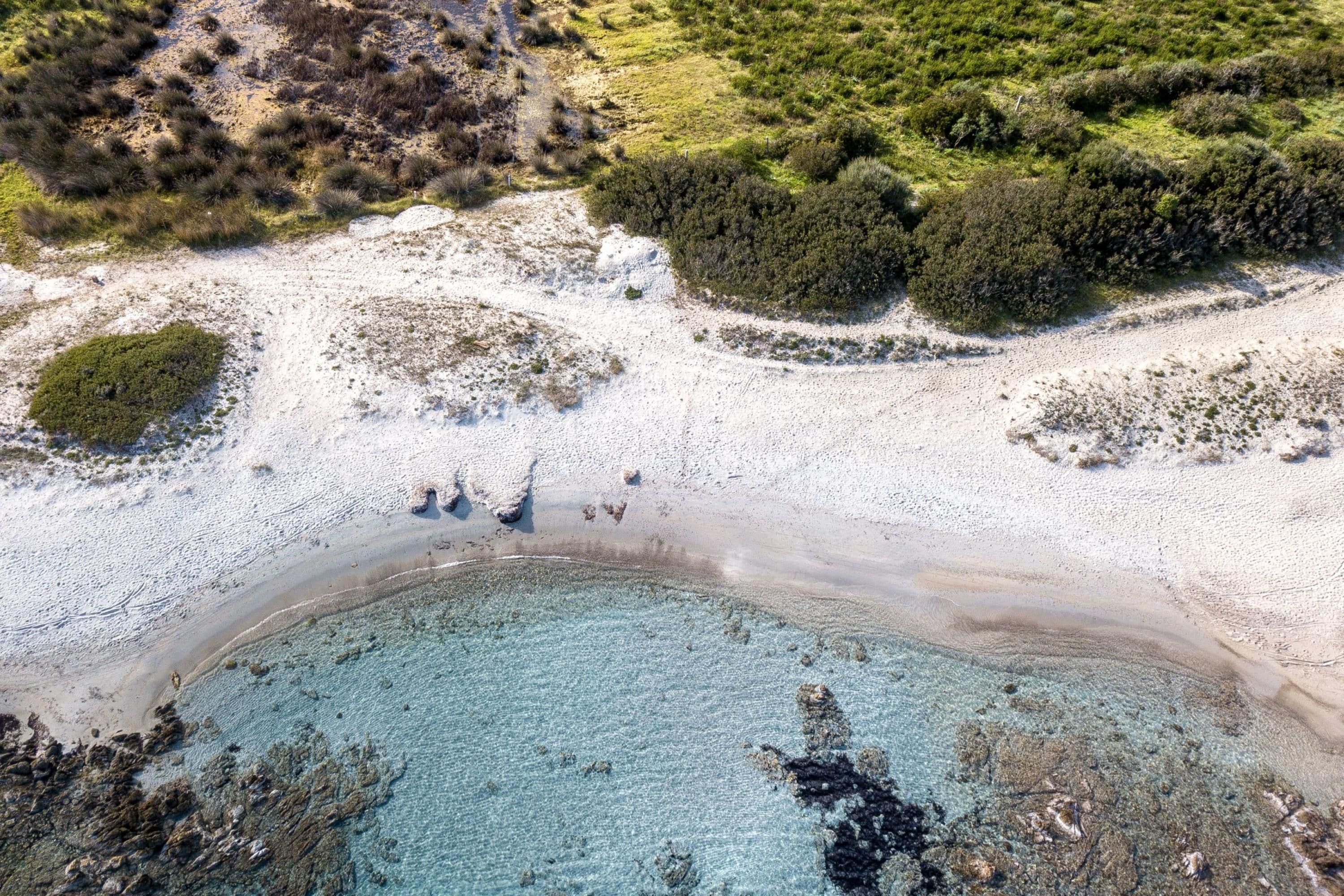 An aerial view of a beach with clear turquoise waters, sandy shores, and sparse vegetation.