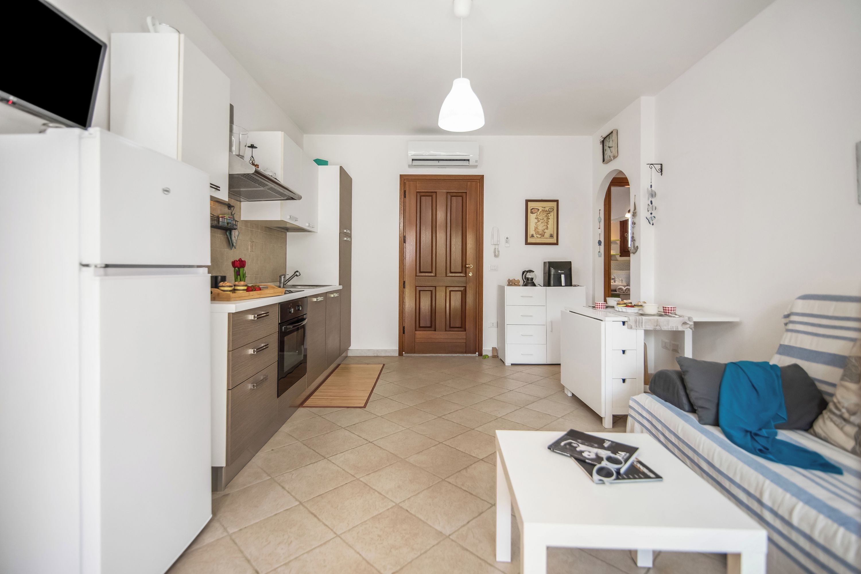 A compact kitchen with a refrigerator, stove, and sink, leading to a wooden door. A white sofa and a coffee table with a magazine are in the living area.