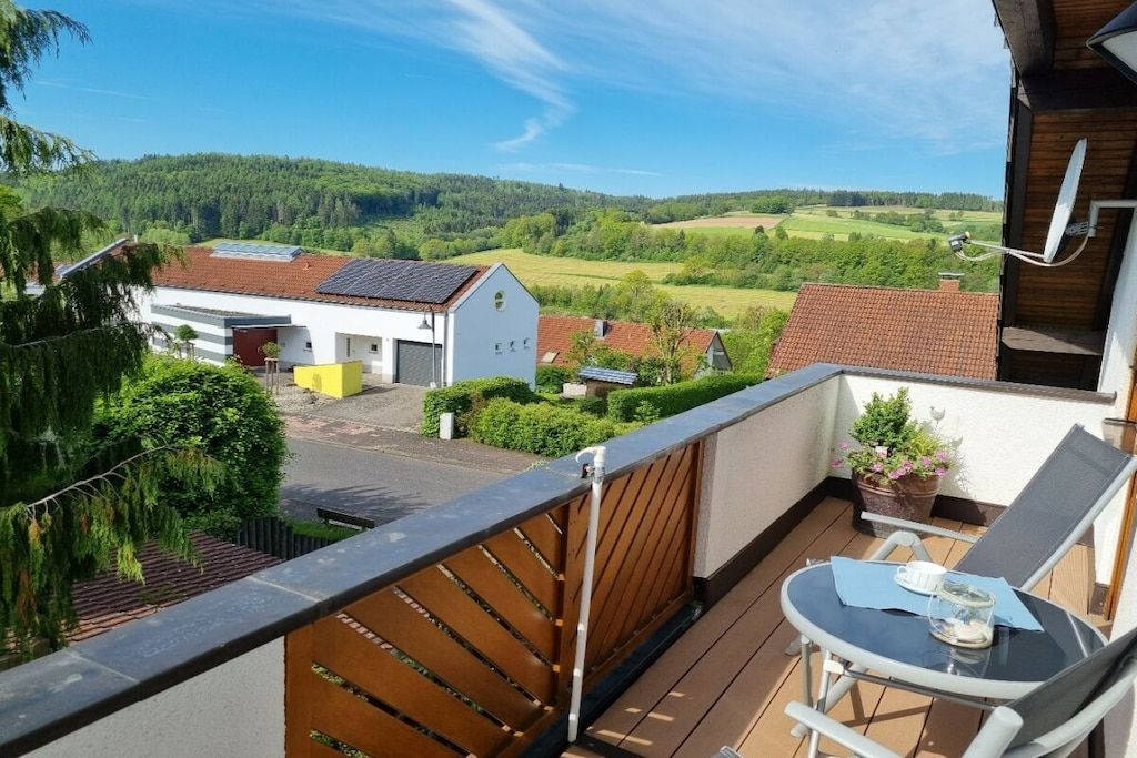 A balcony with a table and chairs, overlooking a residential area with houses and greenery.