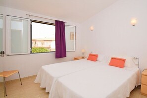 A hotel room with two beds, a window with purple curtains, a small table, and a wall-mounted lamp. - Terraced house with 1 bedroom (Sa Pobla)