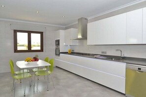 A modern kitchen with white cabinetry, a built-in oven, and a dishwasher. There is a dining area with a white table and green chairs.