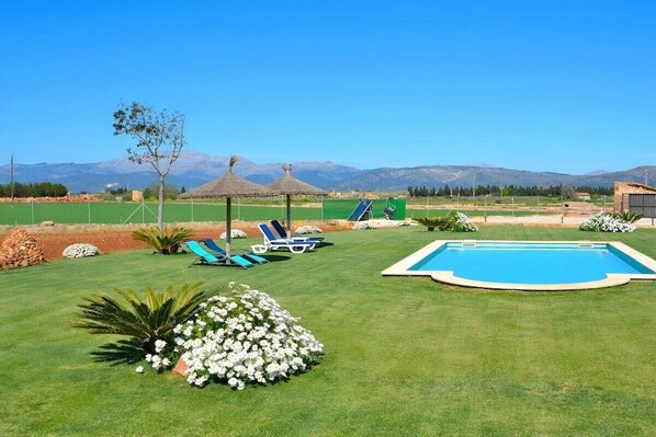 A pool area with a clear view of mountains in the background, surrounded by well-maintained grass and a few lounge chairs.
