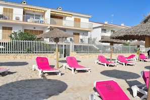 A beachfront property with multiple balconies, a sandy beach area with sun loungers, and thatched umbrellas. - Apartment in Alcudia with terrace (Alcudia)