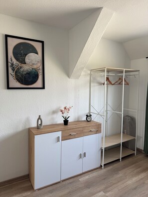 A room with a white wardrobe, a small table with a vase and flowers, a framed picture on the wall, and a white shelving unit with hangers.