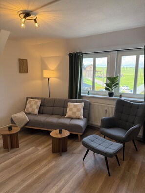 A modern living room with a grey sofa, a grey armchair, a small round table, and a floor lamp. There is a window with a view of a green landscape.