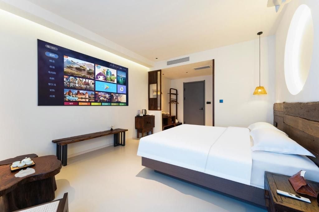 Quarto de hotel moderno com um grande painel digital na parede, uma cama, uma mesinha de cabeceira, um banco e uma pequena escrivaninha com cadeira.