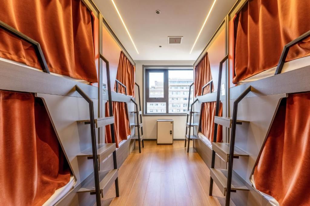 Un pasillo con literas, una ventana con vista a edificios y un refrigerador pequeño.