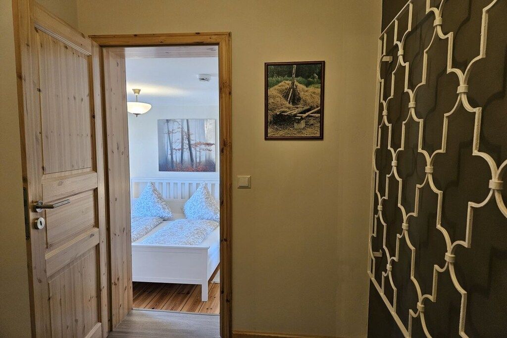 Een gang die leidt naar een slaapkamer met een bed, een ingelijst schilderij aan de muur en een decoratief scherm aan de rechterkant.