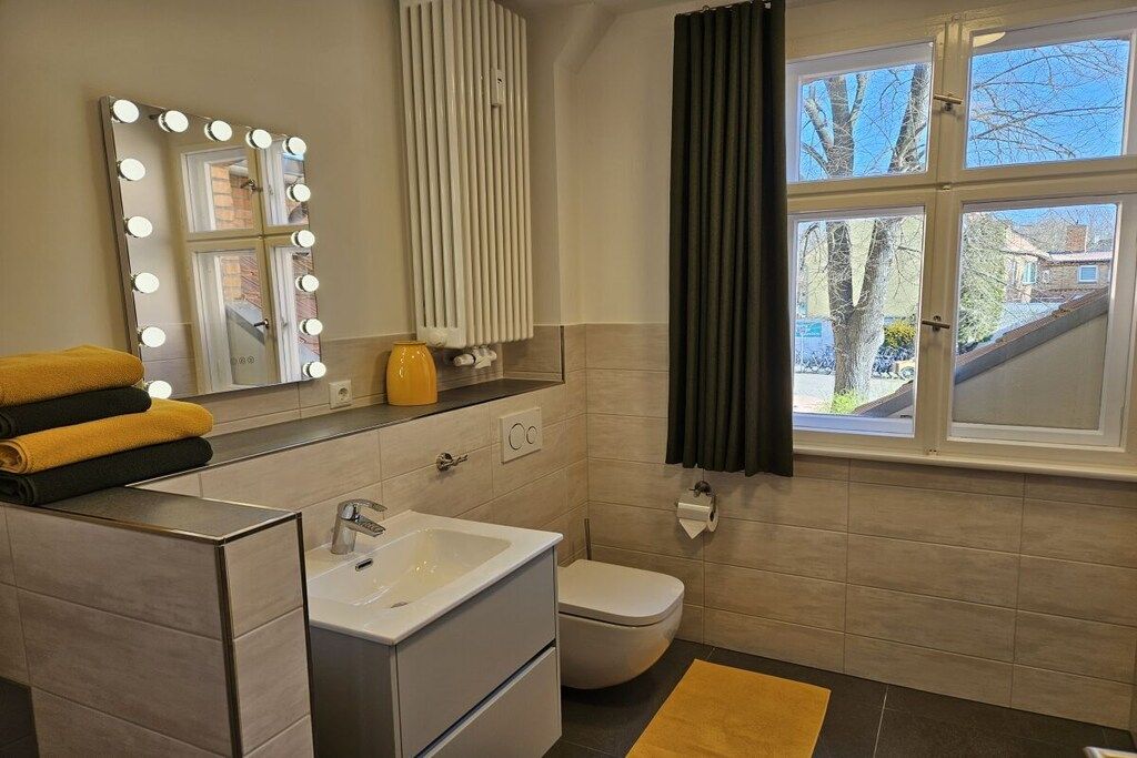Ein Badezimmer mit Waschtisch, Spüle, beleuchtetem Spiegel, Toilette und einem Fenster mit Vorhängen.
