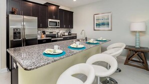A modern kitchen with dark cabinets, stainless steel appliances, a granite countertop, and a bar area with white bar stools. - Go 43 Champions Gate (Davenport)
