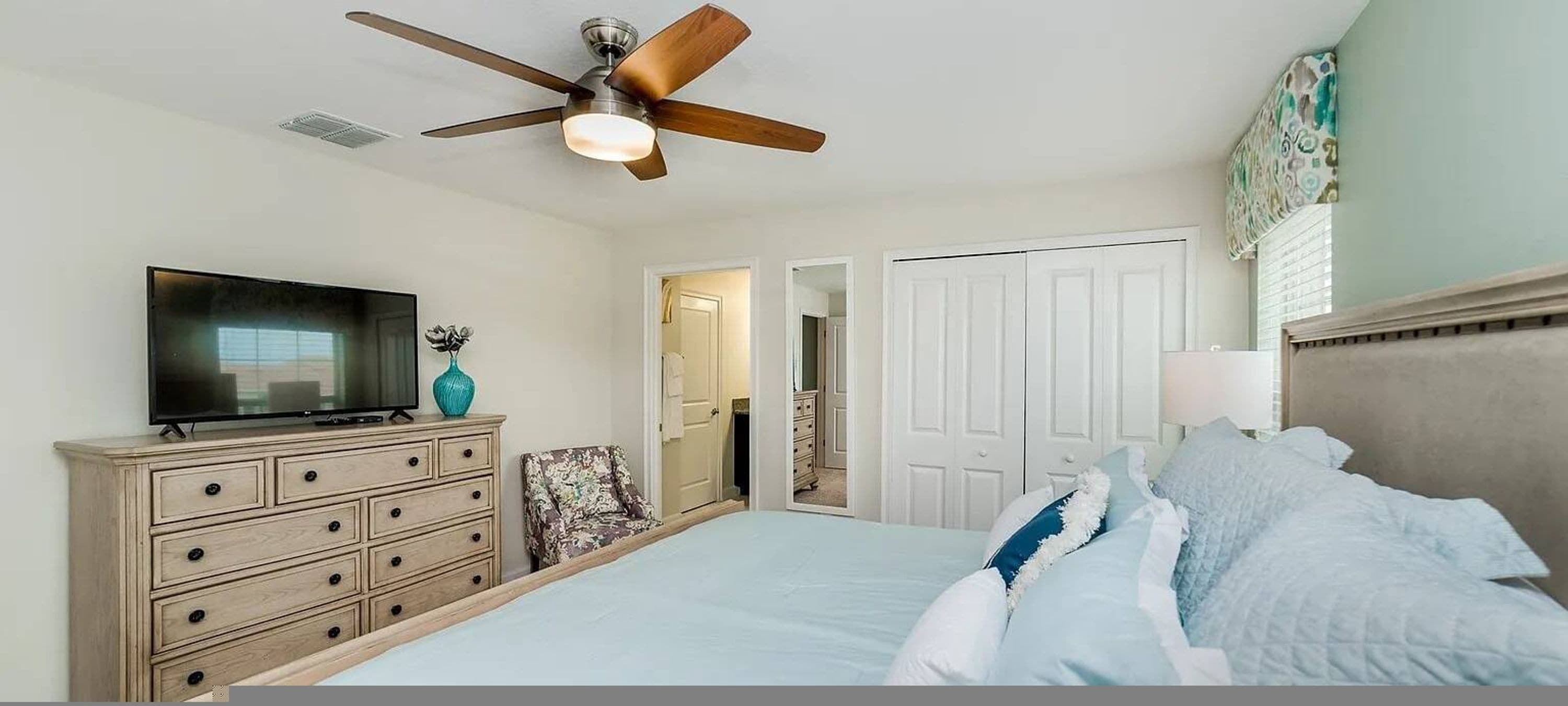 A bedroom with a bed, a dresser, a television, a ceiling fan, and a door leading to another room.