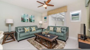 A living room with a sectional sofa, two side tables, a coffee table, a ceiling fan, and artwork on the walls. - Go 43 Champions Gate (Davenport)