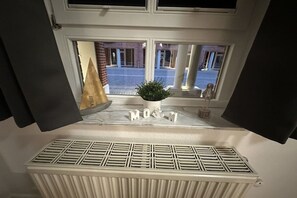 Ein Fenster mit Blick auf ein Gebäude, ein Heizkörper und Dekorationselemente, darunter eine Pflanze und die Buchstaben 'MOM'.