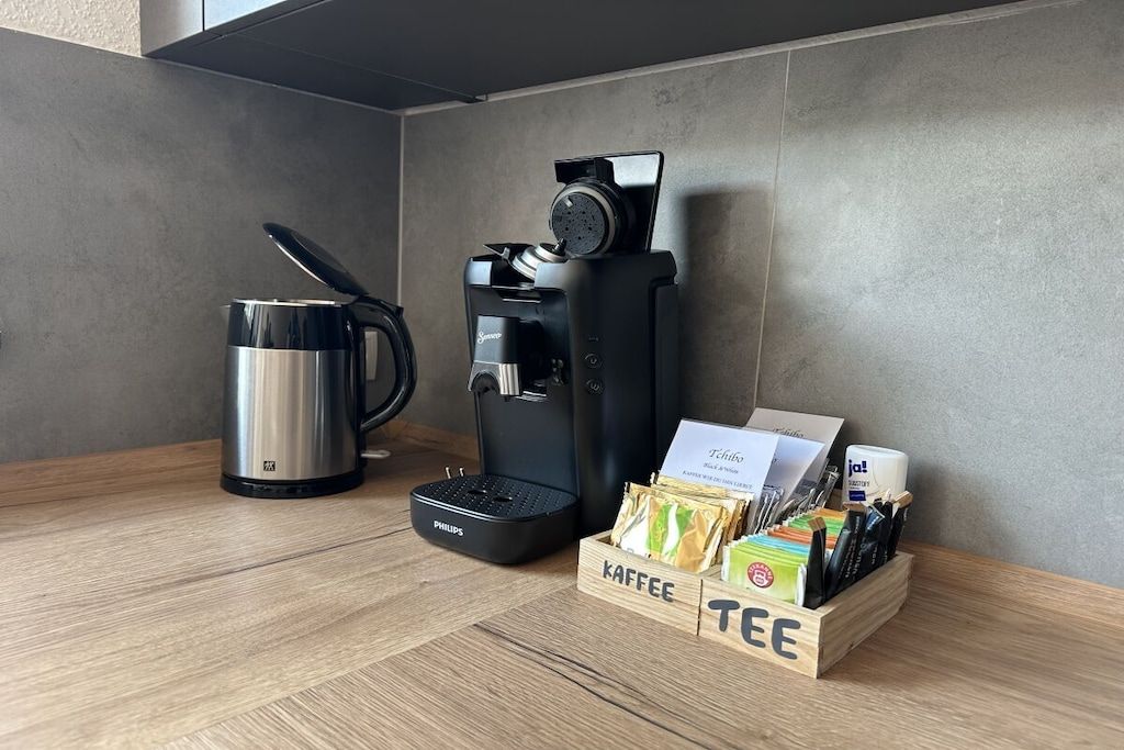 Estação de café com máquina de café, jarra térmica e caixa com o rótulo 'KAFFEE' contendo cápsulas de café e utensílios.