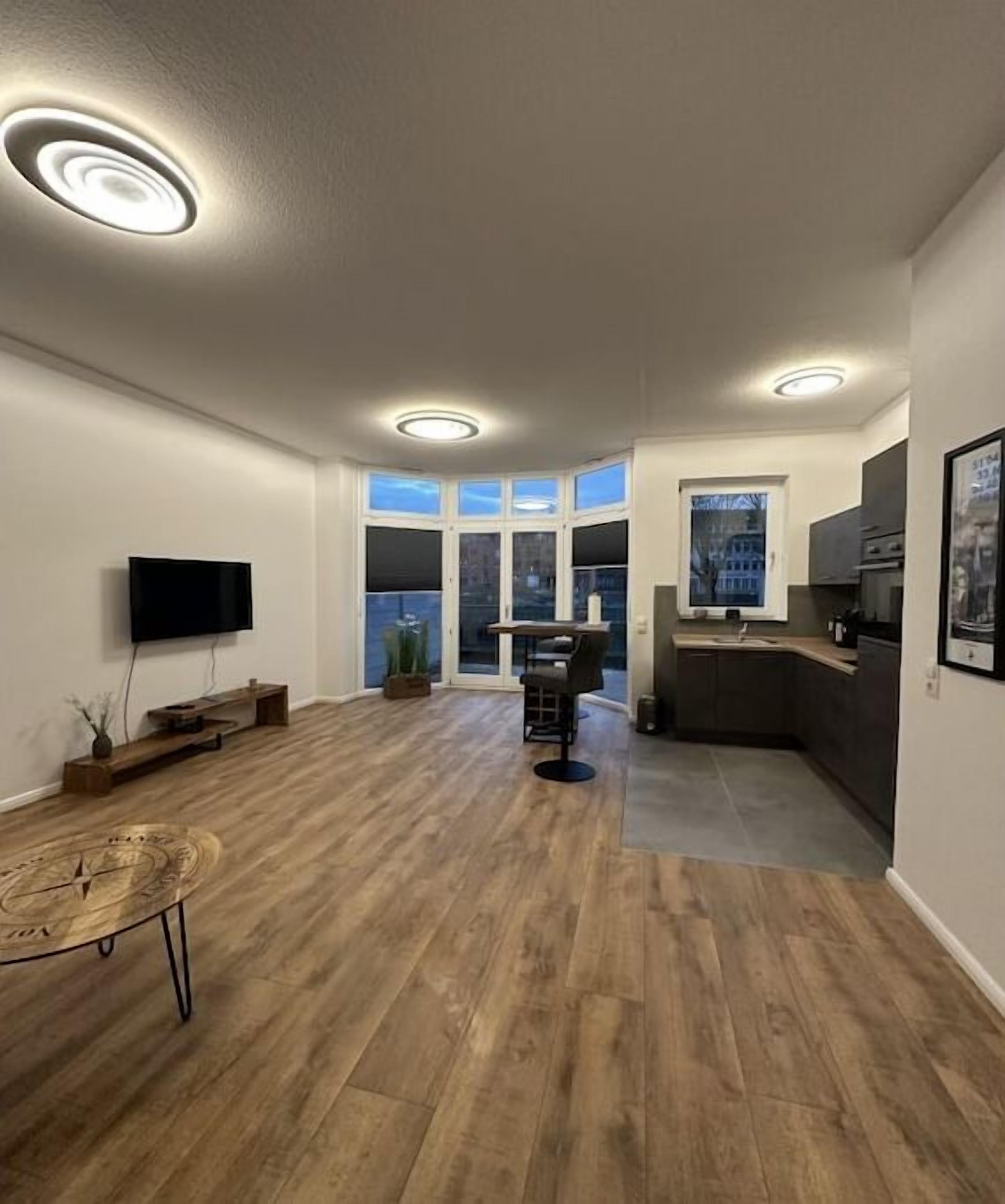 A modern living room with a wooden floor, a flat-screen TV, a dining table, and a kitchen area with a sink and cabinets.