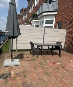 En udendørs terrasse med et bord, stole og en grå parasol, samt en murstensvæg.