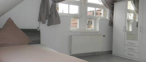 Ein Schlafzimmer mit Bett, Heizkörper, einem Fenster mit Vorhängen und einem Kleiderschrank.