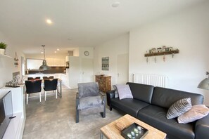 A modern living room with a black leather sofa, grey armchair, and a coffee table. There is a dining area with a table and chairs, and a kitchen in the background. - Bungalow with 1 bedroom (Callantsoog)