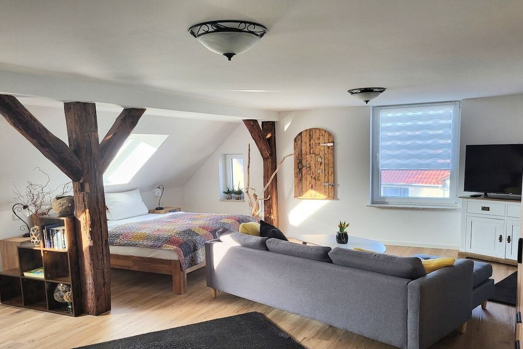 A modern loft bedroom with a sloped ceiling, a large window, a bed, a sofa, and a television.