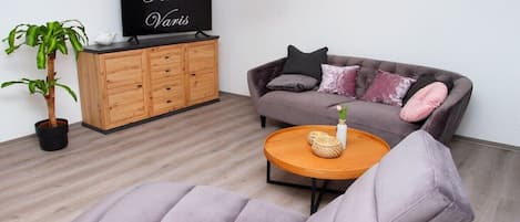 Una sala de estar con un sofá, una mesa de centro, un televisor sobre un mueble de madera y una planta en maceta.