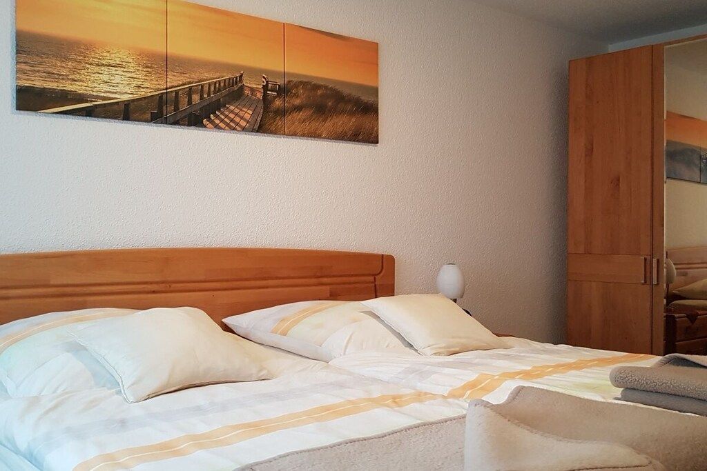 Une chambre à coucher avec une tête de lit en bois, un lit recouvert de linge de lit blanc et un tableau représentant un coucher de soleil sur une jetée.