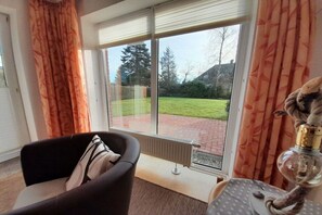 Une pièce dotée d’une porte-fenêtre coulissante donnant sur un jardin, d’un radiateur, d’une chaise et d’une table avec une lampe.