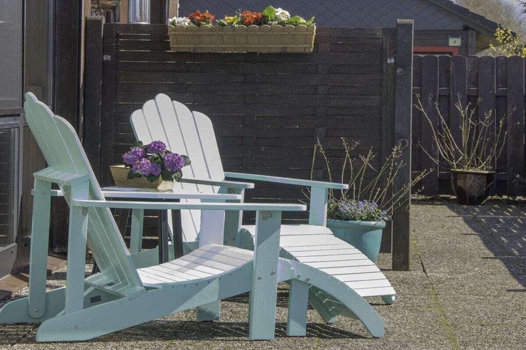 Dos sillas Adirondack con una mesa delantera, una maceta con flores moradas y una cerca de madera al fondo.