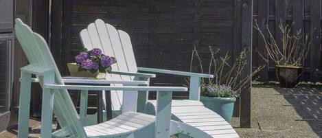 Dos sillas Adirondack con una mesa delantera, una maceta con flores moradas y una cerca de madera al fondo.