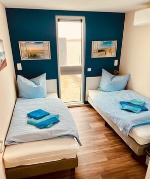 A room with two beds, each with a blue blanket and pillow, a wooden floor, and a window with a view. - Holiday home with 2 bedrooms (Ribnitz-Damgarten)