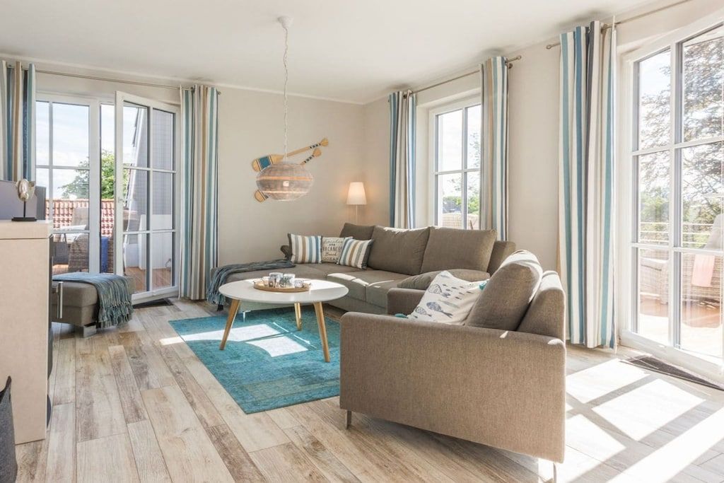 A modern living room with a sofa, a coffee table, and a lamp. There are large windows with curtains, and a view of the outside.