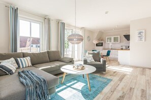 Un salon moderne avec un canapé, un fauteuil et une table basse, une cuisine en arrière-plan et de grandes fenêtres avec des rideaux.