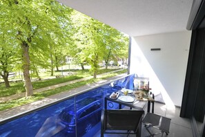Ein Balkon mit Esstisch, Stühlen und Blick auf einen Park mit Bäumen und einem Auto.