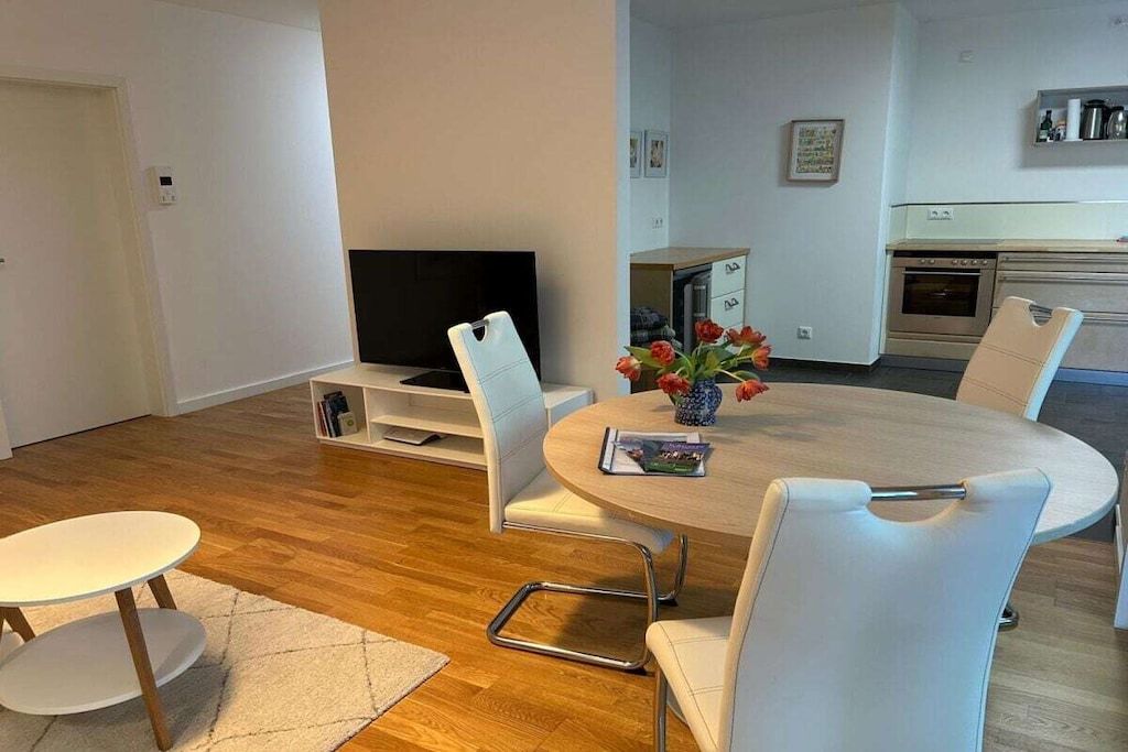 A modern living room with a round dining table, white chairs, a flat-screen TV, and a kitchen area in the background.