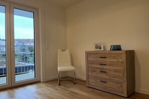 Een kamer met een schuifglasdeur die uitkomt op een balkon, een witte stoel en een houten kast met lades.