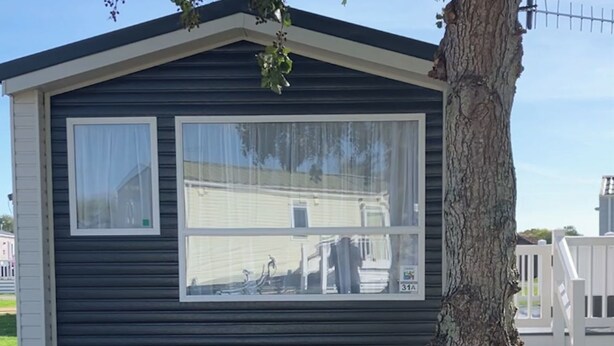 Une maison mobile avec une grande fenêtre, un arbre devant et une clôture blanche en arrière-plan.