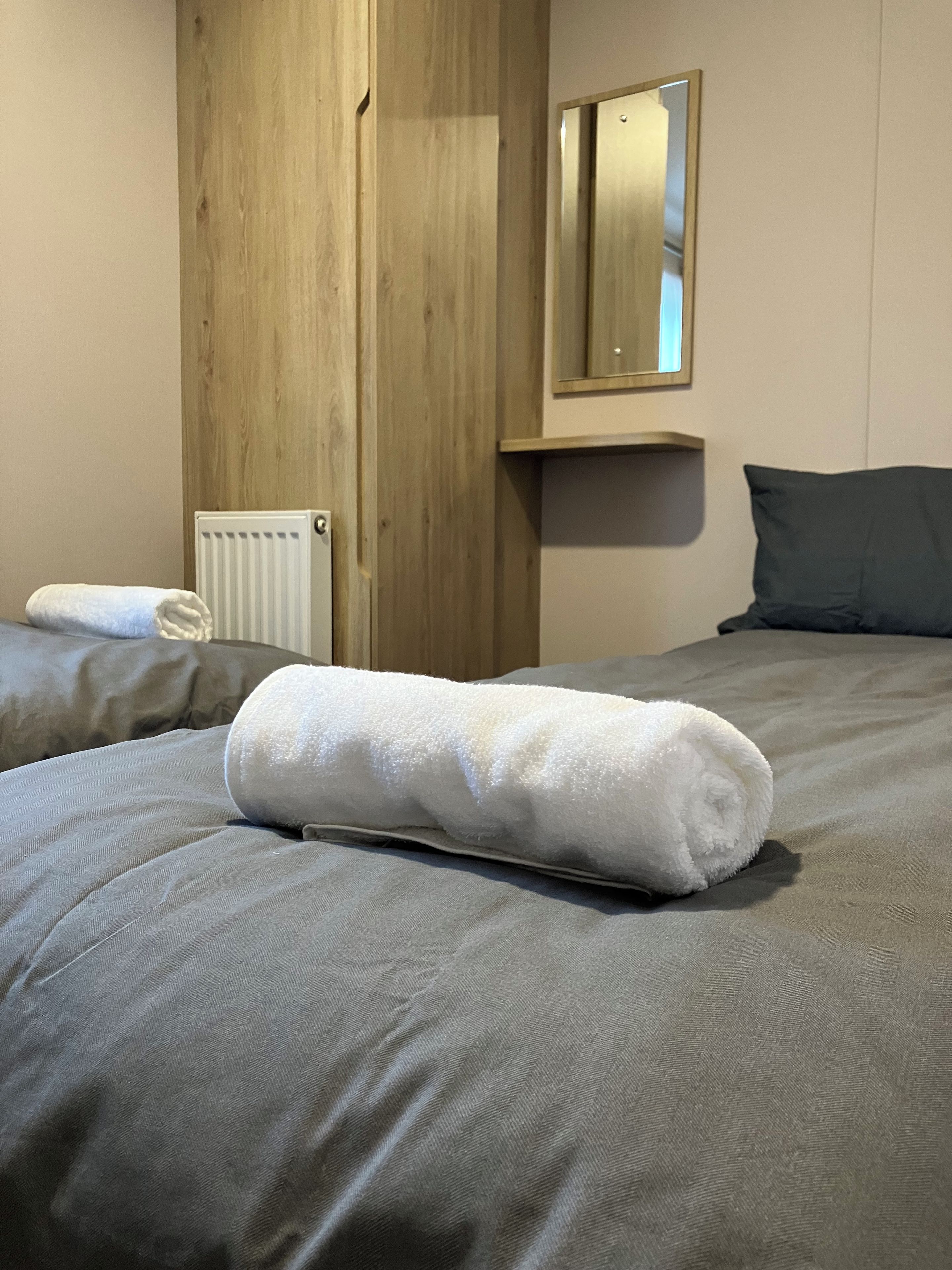 Una cama bien hecha con una toalla blanca enrollada, un armario de madera, un radiador y un espejo en la pared.