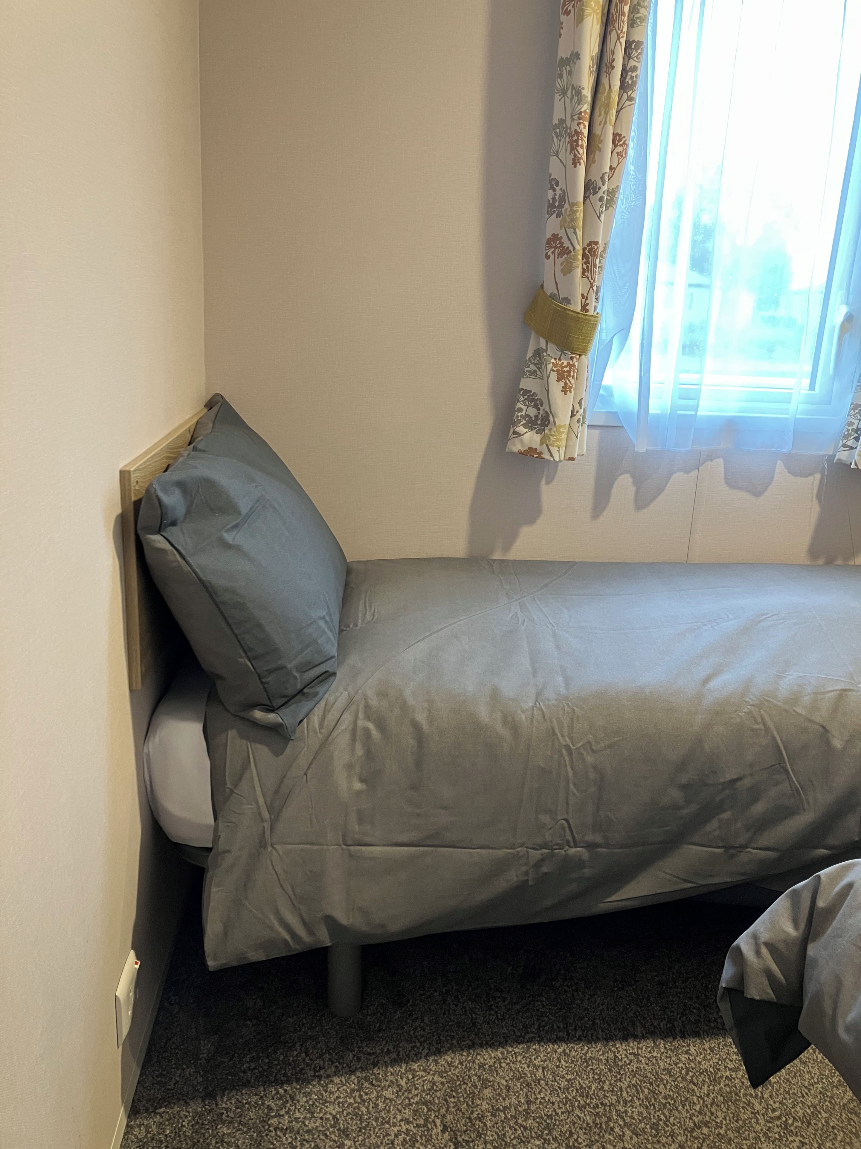 A small, single-bed room with a headboard, a window with floral curtains, and a carpeted floor.