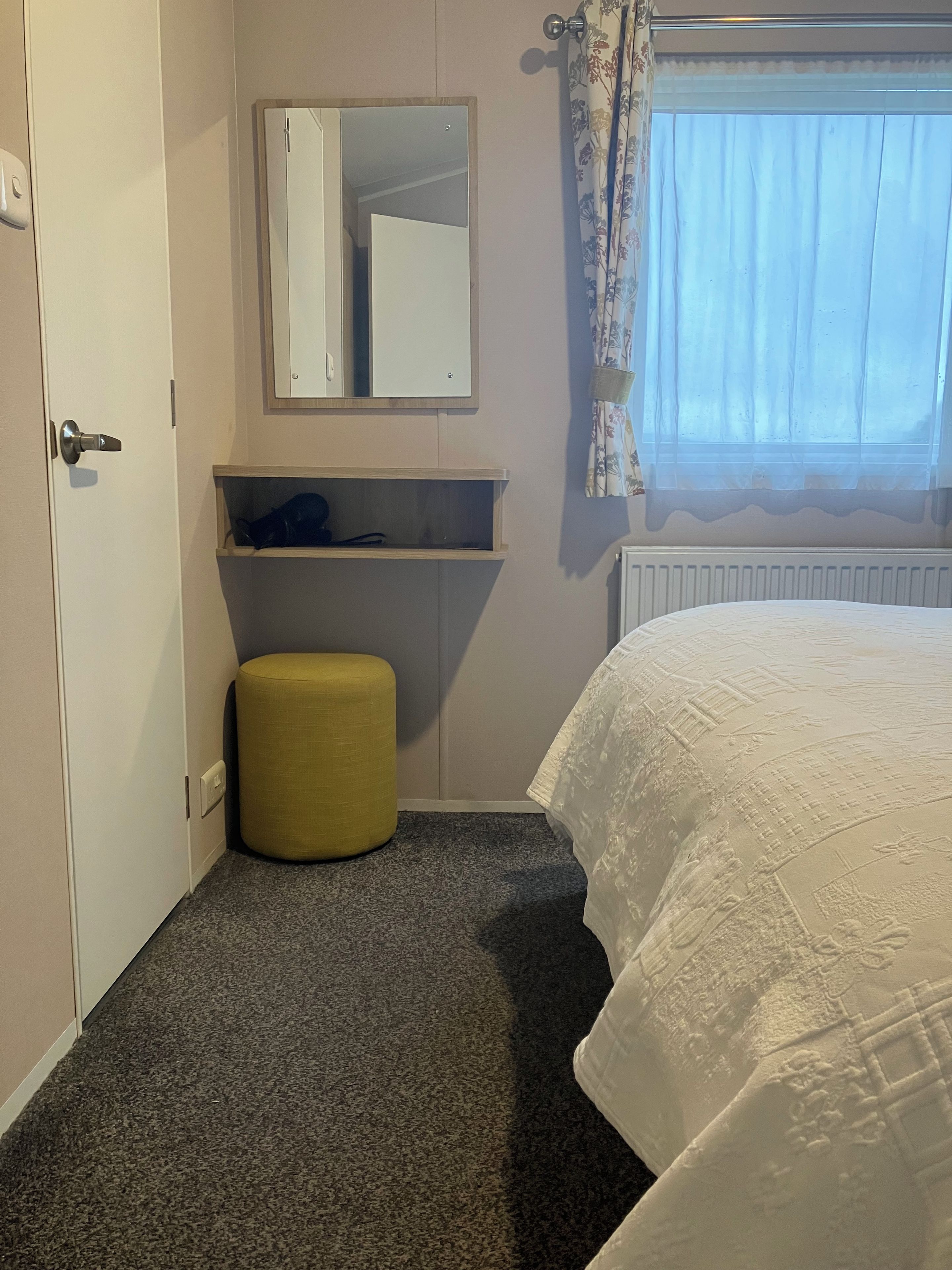 A hotel room with a bed, a small stool, a mirror, and a radiator.