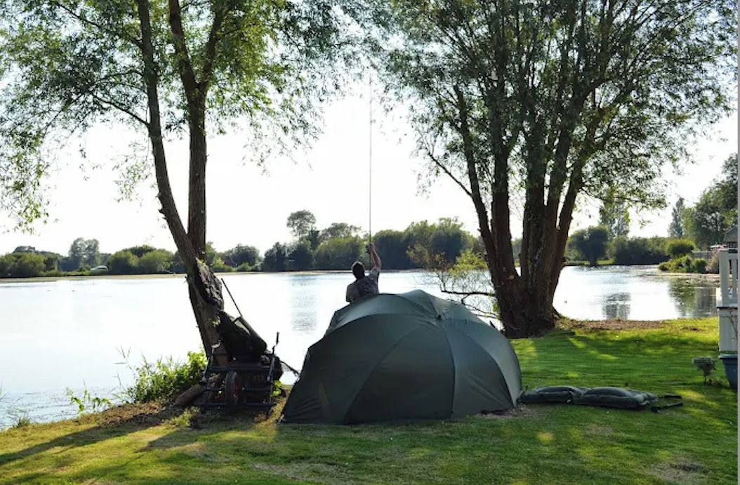 Une tente installée au bord d’un lac, où une personne est en train de pêcher, entourée d’arbres et d’une eau calme.