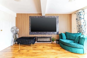 A modern living room with a large flat-screen TV mounted on a wooden wall, a teal sofa, and a wooden floor. - Spacious 4BDR Home Garden and Parking in Windsor (Slough)