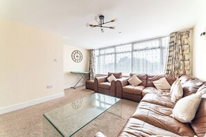 A living room with a brown leather sofa, a glass coffee table, a wall clock, and a large window with curtains. - Spacious 4BDR Home Garden and Parking in Windsor (Slough)