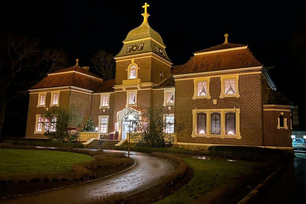 Een groot, bakstenen gebouw met een opvallende toren en een kruis bovenop, met meerdere ramen en een goed verlichte ingang.