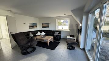 Een moderne woonkamer met een modulaire bank, een schommelstoel, een salontafel en een groot raam met uitzicht op de omgeving.
