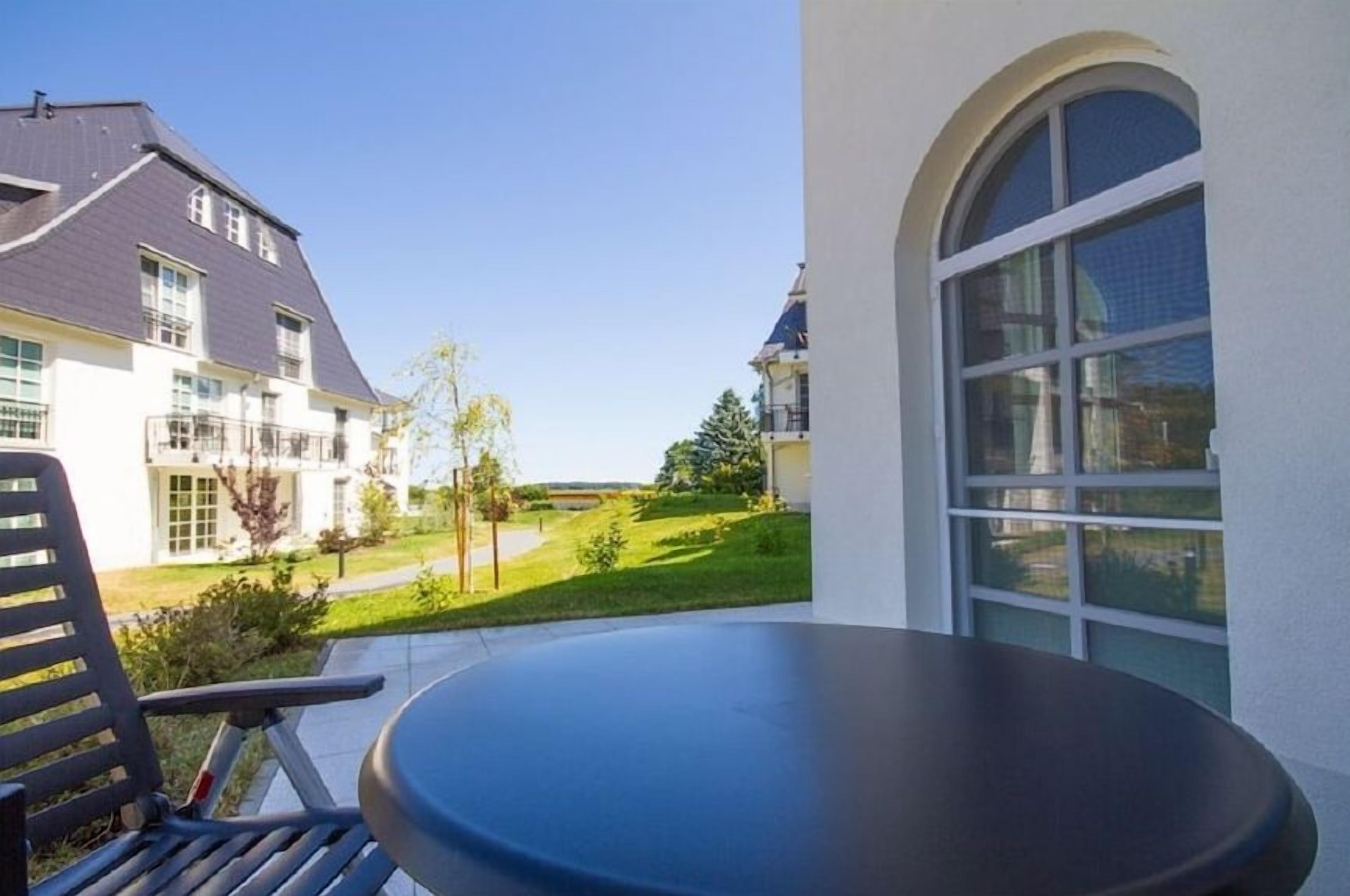 A balcony with a table and a chair, overlooking a well-maintained lawn and a building with multiple windows.